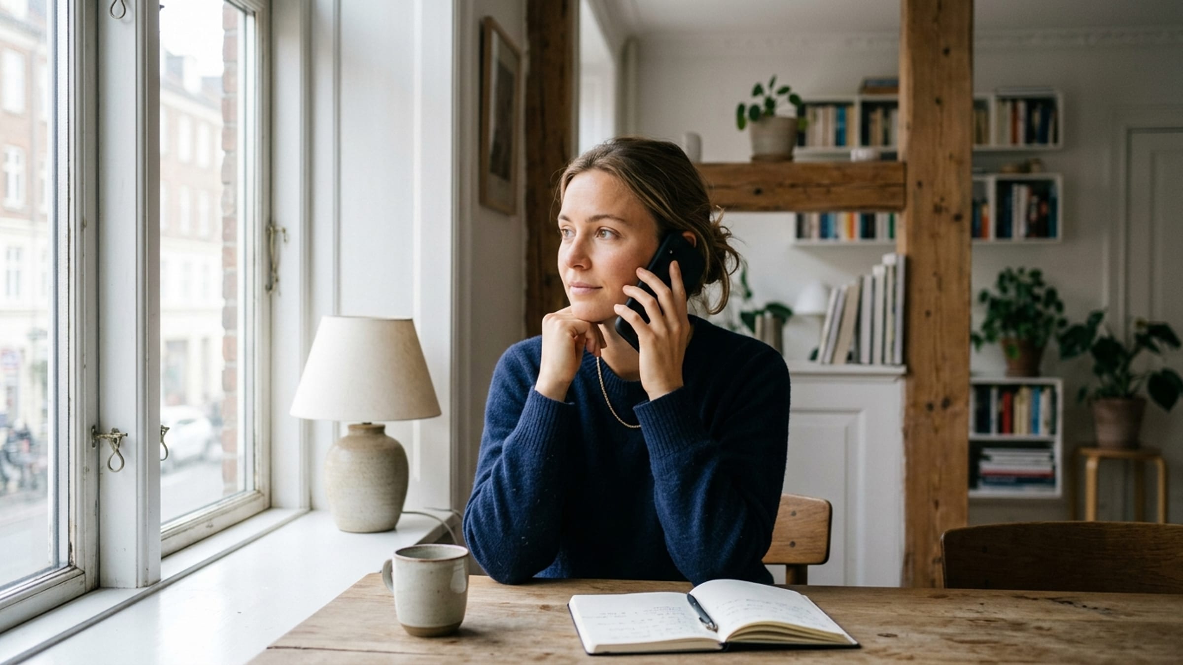 A person seated by a window holding a phone to their ear, listening attentively at the start of a call.