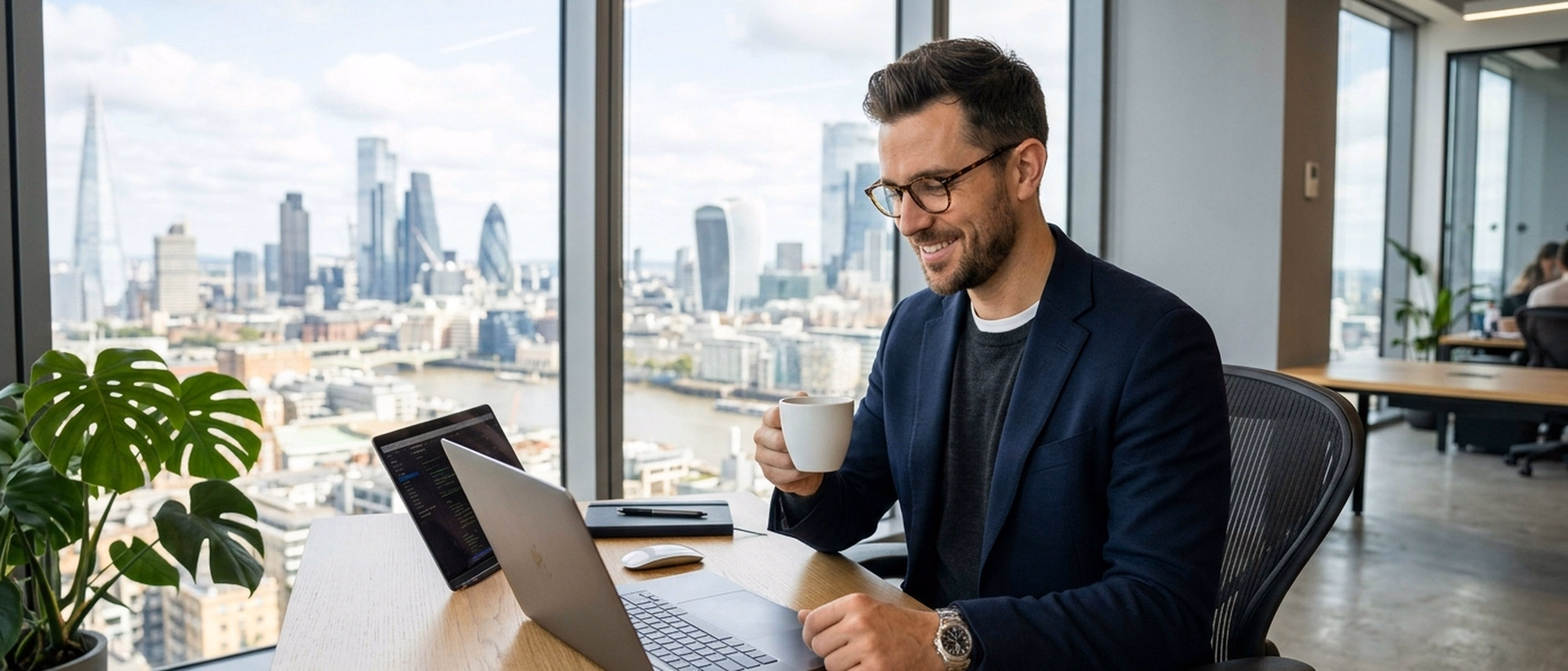 Ein entspannter Experte mit Brille an seinem Laptop mit einer Kaffeetasse an einem Schreibtisch im Hochhaus mit Blick auf die Skyline.